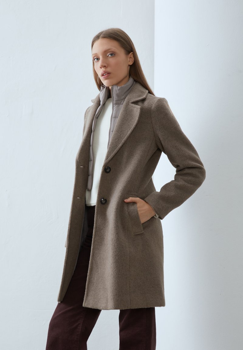 Manteau en laine marron avec un col à revers, doté de deux boutons et de poches latérales. Porté par-dessus une veste matelassée gris clair et un haut blanc.