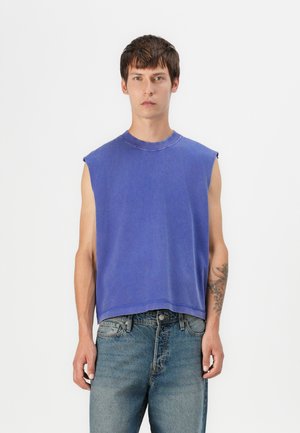 Blue sleeveless top with a round neckline, featuring raw edges and a relaxed fit, paired with light blue denim jeans.