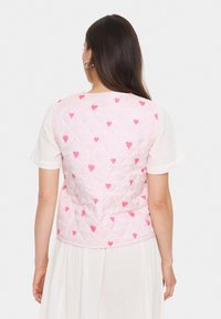 Roza prešita brezrokavnik z diamantskim vzorcem, ki vsebuje majhne rdeče srčke. Brezrokavnik nima vidnih kovinskih delov in je oblečen čez belo srajco.