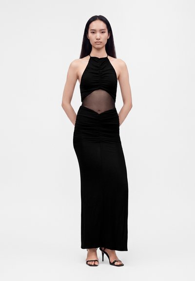 Femme portant une longue robe noire dos nu avec un détail transparent sur la taille, debout les mains dans le dos et portant des talons ouverts noirs.