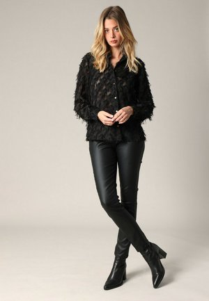 Femme blonde se tient les jambes croisées, portant un chemisier noir transparent texturé, un pantalon en cuir noir et des bottines à talons noirs, sur un fond neutre.