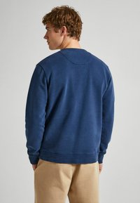 Tumši zila kapuce, kurai ir apaļa kakla izgriezne, raglāna piedurknes un maiga tekstūra, ar teksturētām apmalēm un vienkāršu, gludu dizainu.