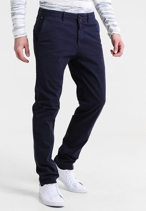 Mężczyzna w ciemnogranatowych spodniach typu slim fit, białych trampkach i szaro-białej koszuli w paski z długim rękawem, stojący z nogami lekko rozstawionymi.