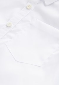 Gros plan de l'avant d'une chemise blanche présentant deux boutons blancs et une poche poitrine carrée sur un tissu lisse.