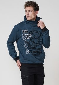 Sudadera azul con patrón y cuello alto, presenta diseños gráficos y texto en negro. Puños y dobladillo acanalados, combinada con pantalones cargo negros.
