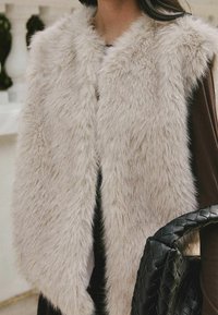 Gilet en fourrure beige et duveteuse porté sur un haut à manches longues foncé, avec un sac à main en cuir noir tressé tenu sur le côté.