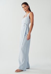 Ženska stoji v svetlo modrem brezrokavnem kombinezonu s pasom in bež sandali na enobarvni beli podlagi.
