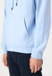 Lys blå bomull hettegenser med kengurulomme, ribbestrikkede mansjetter og snor. Brukt over mørkeblå jeans som har en jevn tekstur.