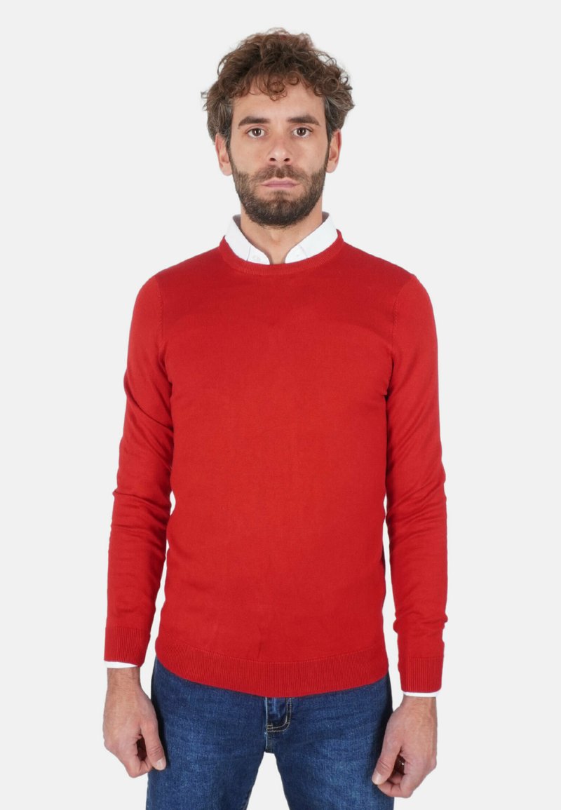 Maglione rosso lavorato a maglia con scollo rotondo, maniche lunghe e polsini e orlo a coste, indossato sopra una camicia bianca con colletto e abbinato a jeans blu.