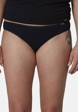 Frau trägt schwarzen Bikini-Unterwäsche und schwarzes Oberteil, zeigt ein Tattoo am oberen Oberschenkel in der Nähe der Hüfte.