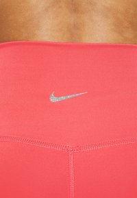 Różowy top sportowy wykonany z gładkiego materiału, z srebrnym, odblaskowym logo Nike w pobliżu dekoltu oraz płaskimi szwami dla komfortu.