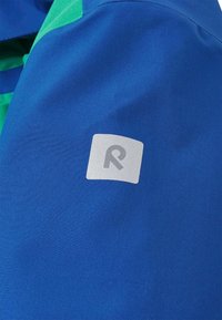 Niebieska tkanina z zielonymi akcentami ma kwadratową metkę w rogu, na której znajduje się szare logo 'R' na białym tle.