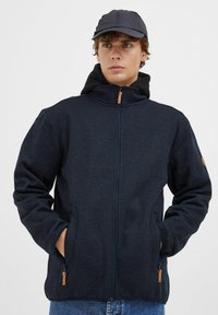Mørk marineblå zip-jakke med en myk teksturert finish, som har sidelommer og hette. Detaljer inkluderer en tan glidelås-feste.