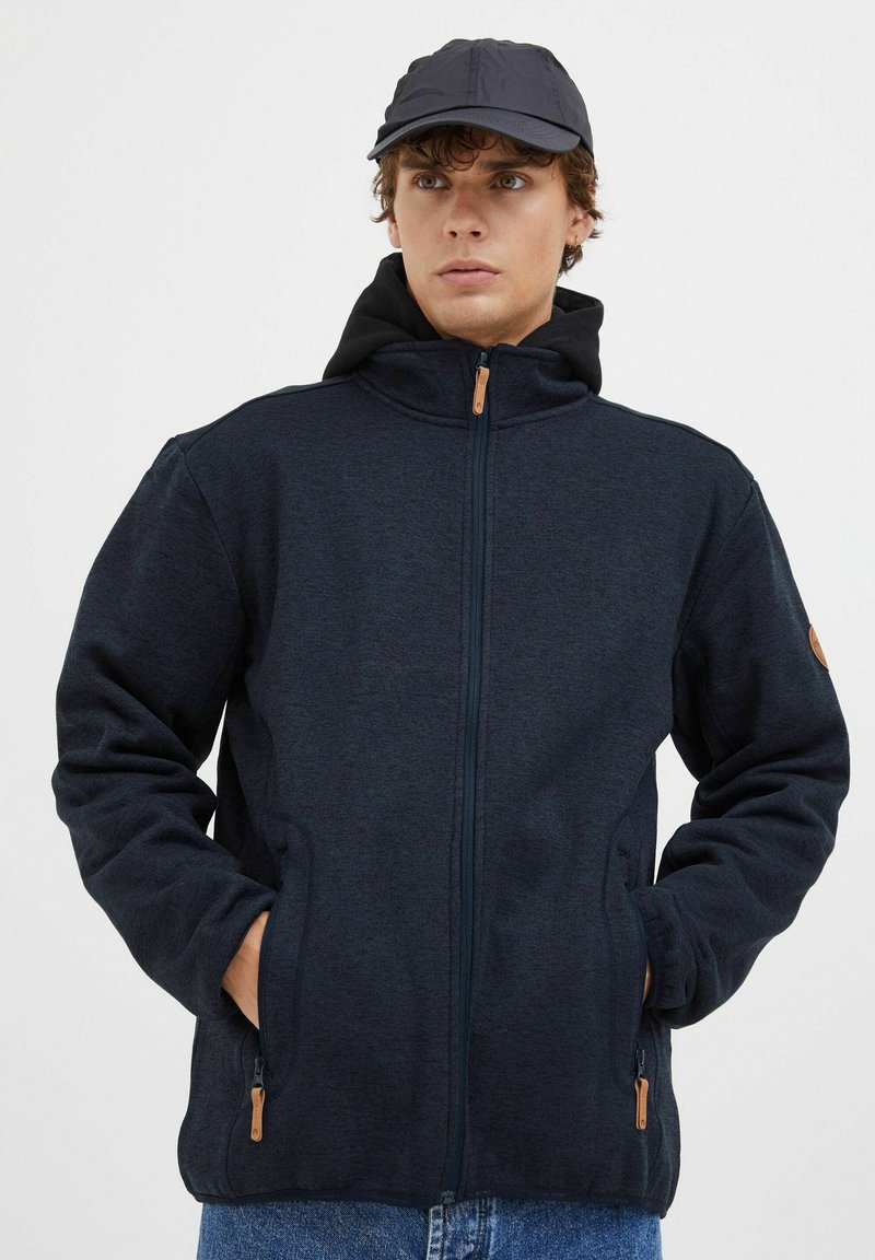 Mørk marineblå zip-jakke med en myk teksturert finish, som har sidelommer og hette. Detaljer inkluderer en tan glidelås-feste.