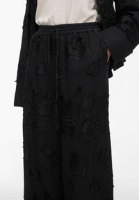Schwarze weit geschnittene Hose mit elastischem Bund und blumiger, strukturierter Stickerei, kombiniert mit einer passenden strukturierten schwarzen Jacke und einem hellbeigen Oberteil.