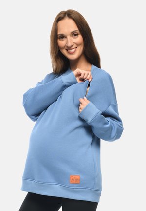 Helles blaues Umstandssweatshirt aus weichem Material, mit einer Reißverschlusstasche und einem kleinen orangefarbenen Logopatch am Saum. Lange Ärmel und lockere Passform.