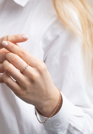 Guldtonad ring med färgglada ädelstenar, bärs på en hand mot en vit skjorta. Slät yta med en delikat design.