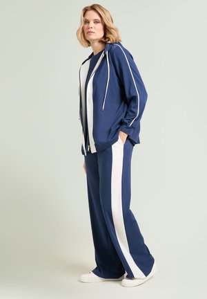 Frau trägt einen marineblauen Trainingsanzug mit weißen Streifen, die Hände in den Taschen, steht mit weißen Turnschuhen vor einem schlichten hellen Hintergrund.