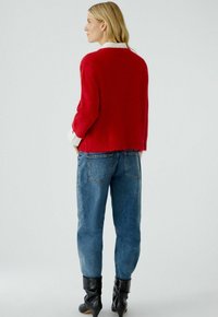 Roter Strickpullover mit lockerem Schnitt und geripptem Saum, kombiniert mit blauen Denim-Jeans und schwarzen Stiefeletten. Schmale Ärmel, sichtbarer Basi-Kragen.
