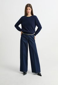 Granatowy sweter z miękkiego materiału, okrągłym dekoltem i kontrastowym jasnym paskiem na hemie, połączony z szerokimi ciemnoniebieskimi dżinsami oraz czarnymi szpilkami.