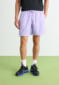 Lyse lilla badebukser med elastisk talje og snøre. Sammen med sorte sneakers med blå accenter og hvide sokker.