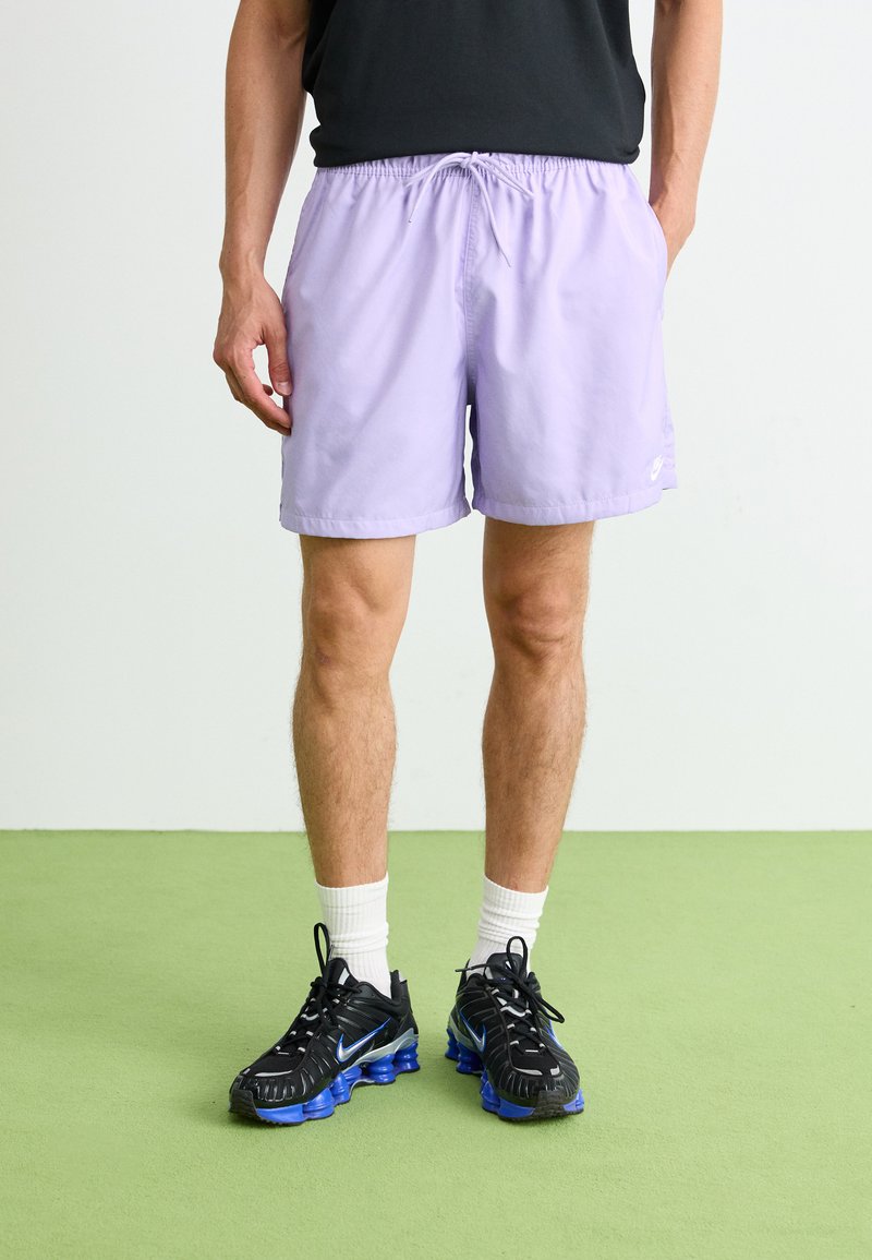 Lyse lilla badebukser med elastisk talje og snøre. Sammen med sorte sneakers med blå accenter og hvide sokker.