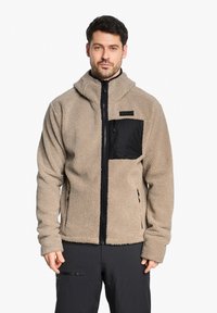 Veste en polaire beige avec une poche poitrine zippée en nylon noir, dotée d'un col montant et de poches latérales, d'une texture douce et d'une coupe décontractée.