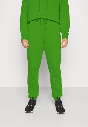 Partie inférieure du corps portant un pantalon de survêtement vert vif et un sweat assorti avec des baskets noires sur un fond blanc uni.