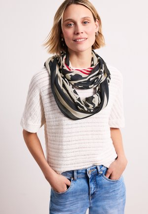 Young woman with short blonde hair wears a white textured short-sleeve top, blue jeans, and a black, beige, and red patterned scarf.