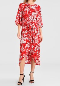 Robe maxi rouge avec des motifs floraux blancs, manches kimono courtes, ceinture à nouer à la taille et fentes sur les côtés. Associée à des talons à lanières noirs.
