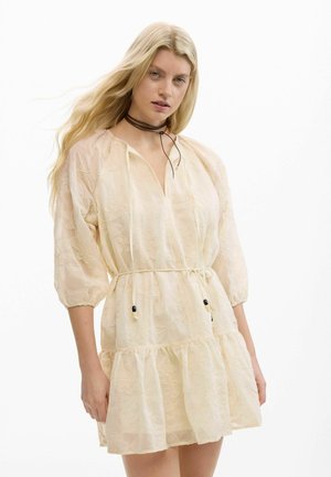 Blondynka w kremowej sukience w kwiaty z rękawami do łokci i cienkim paskiem, uzupełniona brązowym naszyjnikiem typu choker.