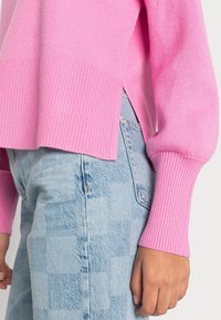 Różowy sweter z dzianiny z żebrowanymi mankietami i dołem, zestawiony z wyblakłymi jasnoniebieskimi dżinsami z geometrycznym wzorem. Sweter ma boczne rozcięcia.