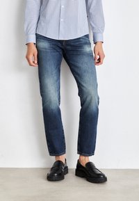 Person med urblekta blå jeans, svarta läderloafers och en blå-vit rutmönstrad skjorta, stående mot en enkel vit vägg.