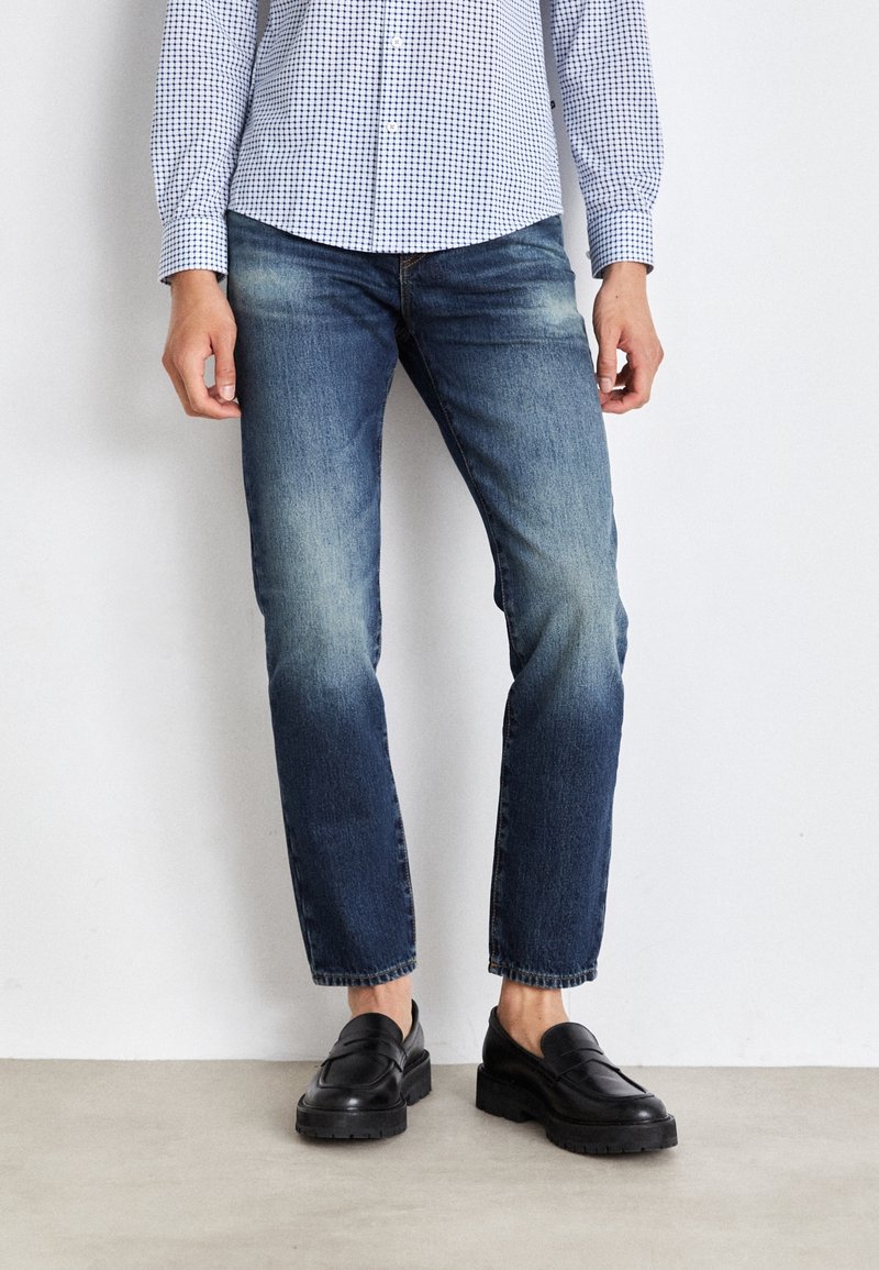 Person med urblekta blå jeans, svarta läderloafers och en blå-vit rutmönstrad skjorta, stående mot en enkel vit vägg.