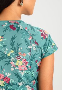 Květinový krátký rukáv top v tazovém materiálu, zdobený růžovými, červenými a žlutými květinami s zelenými listy. Hladká textura a zakřivený výstřih.