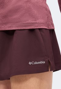 Κοντινό πλάνο ενός ατόμου που φορά βερμούδα Columbia μπορντό και αντίστοιχο μακρυμάνικο μπλουζάκι με υφή, εστιάζοντας στις λεπτομέρειες του ρούχου και το λογότυπο.