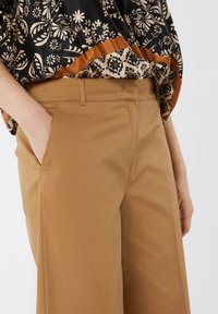 Vaaleanruskeat löysät housut, joissa on sileä rakenne, sivutaskut ja vyötärö. Kuviollinen musta ja beige paita on päällekkäin vyötärön kohdalla.