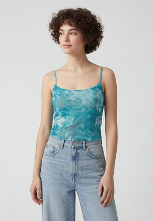Femeie tânără cu păr scurt și creț, purtând un maiou transparent albastru cu imprimeu floral și blugi bleu cu talie înaltă, stând pe un fundal simplu.