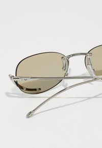 Ochelari de soare cu ramă metalică, lentile maro și o formă elegantă, ovală. Rama prezintă lucrări detaliate din metal și un design curbat al bratelor.
