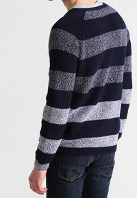 Homme portant un pull en tricot rayé bleu marine et gris clair à manches longues et un jean bleu foncé, tourné de dos à la caméra.