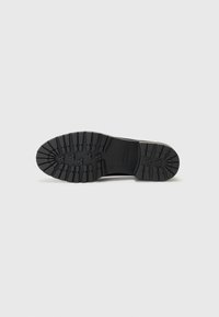 Suela de bota de goma negra con surcos profundos, superficie texturizada y un logotipo rectangular grabado en el centro. Diseño plano y simétrico.