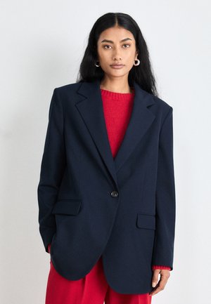 Blazer oversized de marine à la coupe structurée avec un bouton unique, doté de deux poches avant et d'une texture lisse, porté sur un pull rouge.