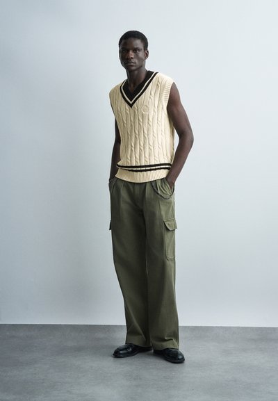 Λεπτός άνδρας στέκεται με τα χέρια στις τσέπες, φορώντας γιλέκο από κρεμ πλεκτό με ανάγλυφα σχέδια χωρίς μανίκια και φαρδιά χακί παντελόνια cargo με μαύρα παπούτσια.