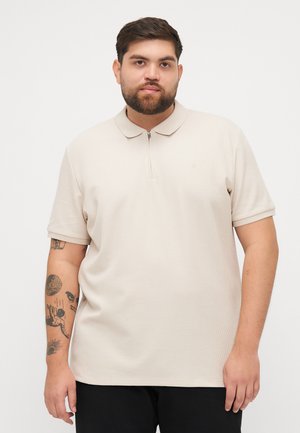 Beige kortærmet polo shirt i tekstureret stof, med lynlås krave og et logo på brystet, parret med sorte bukser.