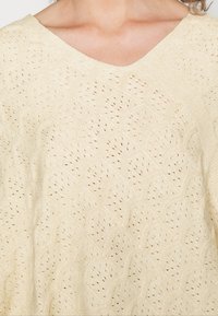 Suéter de malha em bege claro com um padrão texturizado que apresenta formas curvas sobrepostas e um design de pontos abertos. Decote em V.