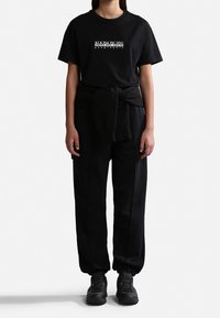 Joven vestido con una camiseta negra de Napapijri, pantalones jogger negros, zapatillas negras y un sudadera negra atada a la cintura, sobre un fondo blanco.