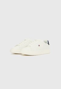 Par belih nizkih superg z vezalkami, s majhno rdečo, belo in modro oznako na strani in temnim jezičkom na peti.