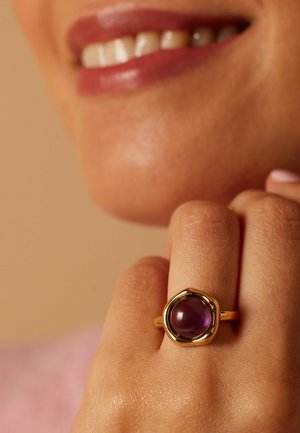 Gros plan sur une main avec une bague en or ornée d'une pierre précieuse ronde violette, près d'une bouche souriante aux lèvres naturelles et aux dents visibles.