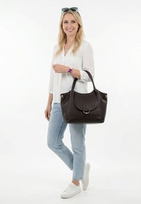 Mujer rubia con blusa blanca, jeans azul claro, zapatillas blancas, reloj morado, gafas de sol en la cabeza, sosteniendo un bolso grande de cuero marrón oscuro.