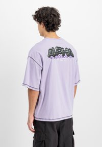 Tricou oversized de culoare mov deschis, cu grafic�ă neagră "ALPHA" și text mov pe spate, cu detalii vizibile de cusătură și mâneci scurte.